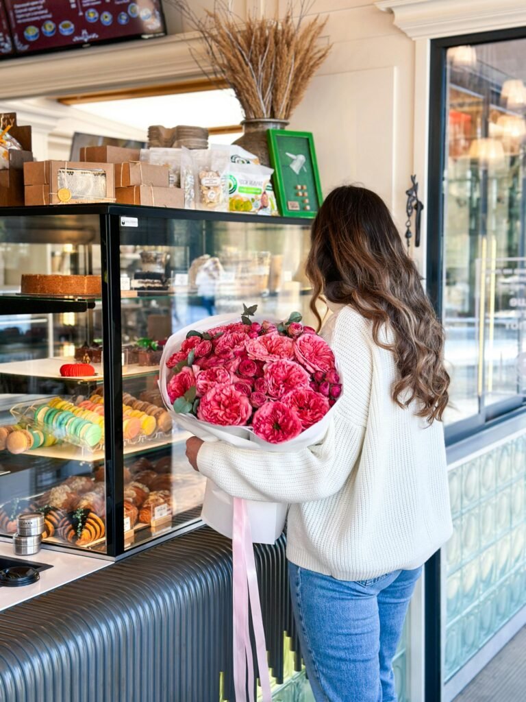 pexels-photo-30839447-30839447 A woman in a cozy sweater holds pink flowers inside a bakery with glass display.